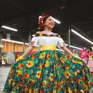 Person in a colorful folkloric outfit featuring a white off-shoulder blouse and a full green skirt with large yellow and red floral patterns, standing indoors and holding the skirt out to display its design.