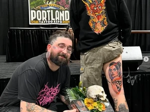 A photo of a tattoo artist with a prize next to the tattoo he did on a persons leg. 