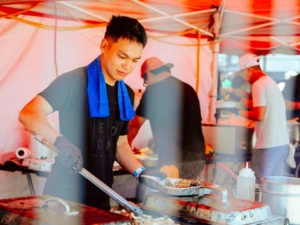 Person cooking at a food stall under a red canopy, using a spatula to prepare food on a large flat-top grill surrounded by trays and cooking supplies.