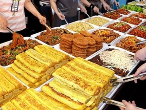 Large buffet-style spread of assorted dishes including stacks of golden fried rolls, noodles, patties, and various stir-fried items, with people serving food using tongs.