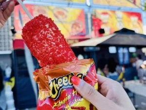 Hand holding a corn on the cob coated in crushed Flamin’ Hot Cheetos, with the Cheetos snack bag used as a holder, in front of colorful food stand signs outdoors.