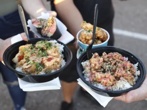 Three hands holding bowls of rice topped with poke, crispy fried pieces and garnished with herbs and sauces, along with a sushi roll in a cup.