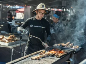 Person grilling meat bones over an open flame at an outdoor food stall, using a blowtorch to sear the meat, with smoke rising from the grill and trays of bones nearby.