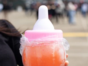 Person holding a large baby bottle-shaped container filled with bright red and orange beverage, wrapped in a plastic bag, outdoors.