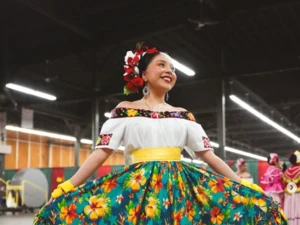 Person in a colorful folkloric outfit featuring a white off-shoulder blouse and a full green skirt with large yellow and red floral patterns, standing indoors and holding the skirt out to display its design.