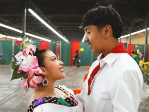 Two people dancing in traditional attire indoors; one wears a white shirt with a red scarf, and the other wears a vibrant dress with floral patterns and large pink flowers in the hair.