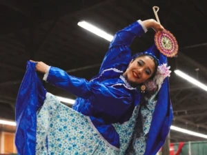 Person wearing a bright blue traditional folkloric dress with a floral-patterned skirt, holding the skirt wide and raising one arm with a decorative fan in an indoor performance space.