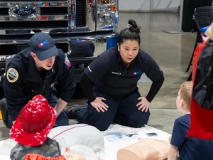 Two EMS workers kneel on the floor demonstrating CPR techniques to young children who sit around practice mannequins. An ambulance is parked behind the instructors.