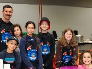 A group of children and one adult wearing aprons and name tags stand behind a table with a red-checkered tablecloth at a kids’ cookoff event. Cups, a voting sign, and supplies sit on the table, and a smiling girl stands in front on the right.