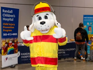 A person in a Sparky the Fire Dog mascot costume wearing a yellow and red firefighter outfit and helmet poses with both thumbs up at an indoor event. A Randall Children’s Hospital booth is visible in the background.