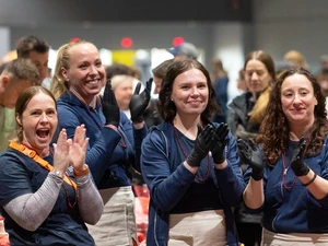 Four women wearing navy shirts, aprons, and black gloves stand behind a table covered with red cloth, smiling and clapping during an indoor community event. Water bottles and cooking supplies sit on the table in front of them.