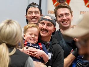 Three smiling firefighters stand behind a laughing young boy who is being held in someone’s arms at a community event. The group is gathered in front of a booth while other attendees move around the background.