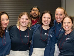 Six adults wearing navy shirts, aprons decorated with horses, and red bead necklaces stand together smiling behind a table with several crockpots and serving supplies. A checkered tablecloth covers the table.