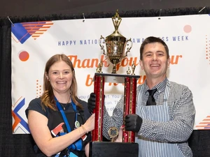 Two adults stand in front of a “Thank you to our burn nurses” banner while holding a large gold and red trophy. Both are smiling; one wears an apron and gloves and the other wears an event badge.