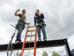 Two women wearing safety harnesses stand on a steel beam above a ladder, flexing their arms.