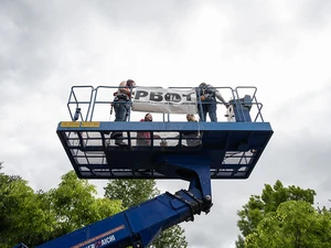 Three people stand in a raised lift platform installing a banner overhead.