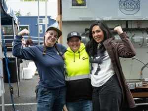 Three women pose together smiling and flexing in front of industrial equipment.