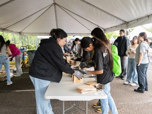 Students practice hands on woodworking skills at a table under a tent while others watch.