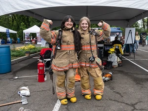 Two girls wearing firefighter gear stand together flexing their arms at an outdoor booth.