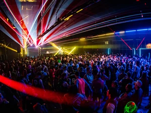 Large audience at an indoor electronic music event, gathered around a stage with a DJ, as bright multicolored lights and beams fill the venue.