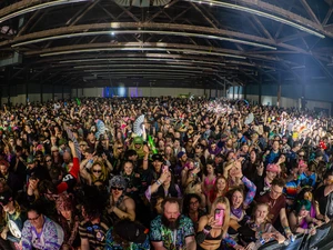 Dense crowd at an indoor concert venue, with attendees facing a stage area and holding up phones and hands under exposed wooden beams.