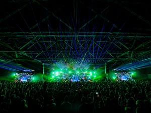Wide view of a packed audience inside a large hall, watching a stage with vivid green lighting, laser effects, and large digital screens.