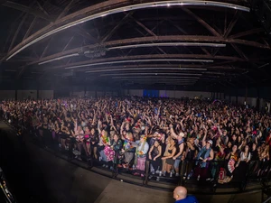Crowded indoor concert scene with attendees lined along a front barrier, many raising hands and phones, beneath a high ceiling with exposed beams.