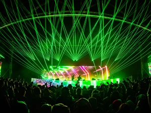 Packed audience at an indoor electronic music show, with raised hands in front of a DJ booth and vivid green laser lights and neon stage visuals overhead.