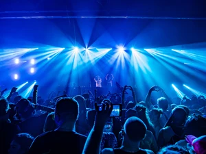 Large audience at an indoor electronic music show, with raised hands and phones in front of a DJ booth, as bright blue lights fan out across the venue.